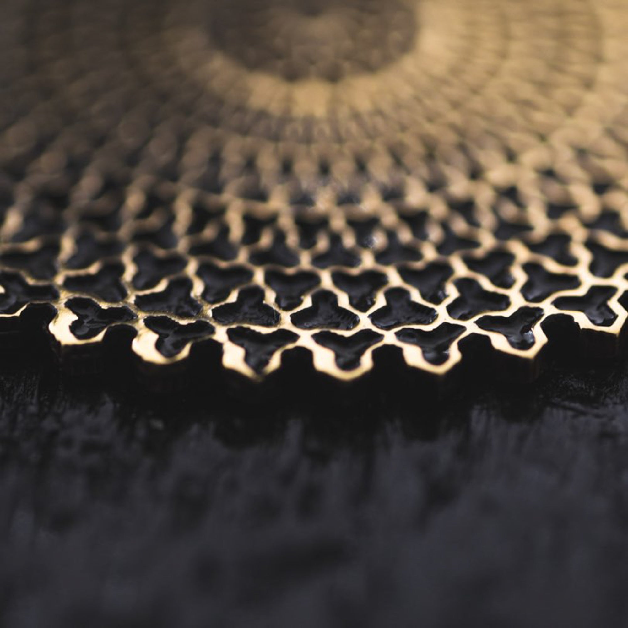 Close-up photo of an intricate gold pattern against a dark background. The design features interconnected geometric shapes, reminiscent of the VORTEX Tribal Oversized Mandala Disc Hoop Earrings in Gold by Kaya Movement, creating a sense of depth and texture. The image is blurred towards the top, drawing focus to the detailed craftsmanship.